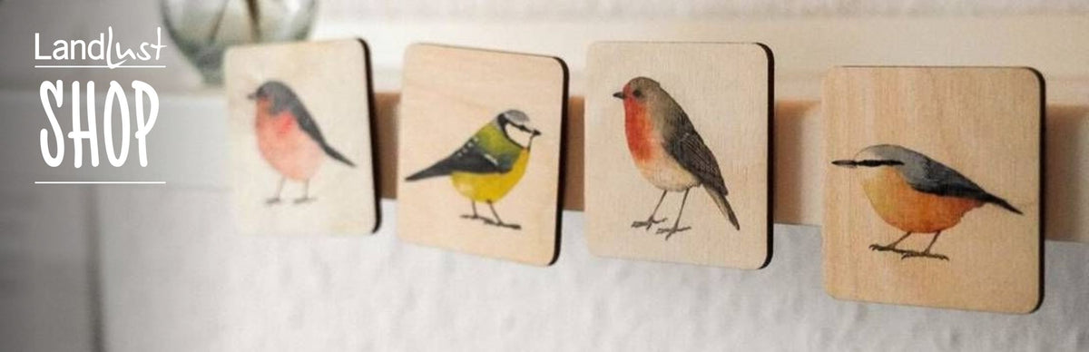 Vier quadratische Holztäfelchen zeigen bunte Vogelillustrationen. Links steht „Landlust Shop“ in weißer Schrift auf grauem Hintergrund.