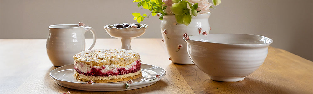 Heller Holztisch mit Sahnetorte, weißen Keramikschalen, Wasserkrug und Blumenstrauß, weiche Farben und zarte Texturen.