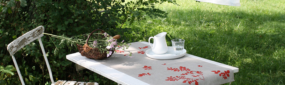 Weißer Holztisch im Garten mit Leinen-Tischdecke, Korb voll Blumen, weißem Krug und Glas Wasser, grüne Wiese im Hintergrund.
