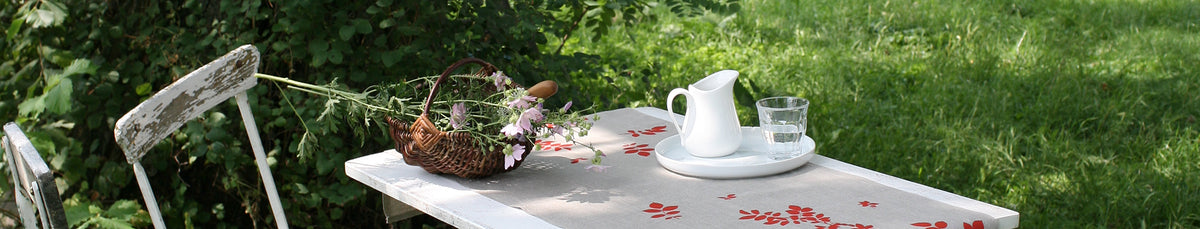 Weißer Holztisch im Garten mit Leinen-Tischdecke, Korb voll Blumen, weißem Krug und Glas Wasser, grüne Wiese im Hintergrund.