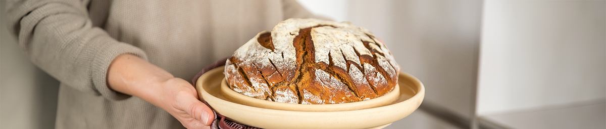 Ein frisches rundes Brot liegt in der Mitte einer cremefarbenen Keramik-Brotbackplatte. Eine Person im beigen Oberteil hält die Platte in der Hand.