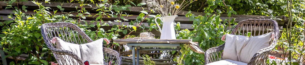 Zwei geflochtene Stühle mit hellen Kissen stehen um einen kleinen Holztisch in einem Garten. Auf dem Tisch befindet sich eine weiße Vase mit Trockenblumen. Hinter der Sitzgruppe sind grüne Pflanzen und ein Holzzaun zu sehen.