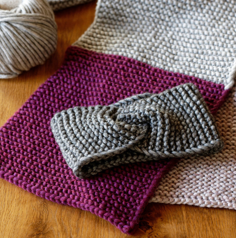 Gestricktes Set aus grauem Stirnband und zweifarbigem Schal in Lila und Grau liegt auf Holztisch neben einem Knäuel hellgrauer Wolle.