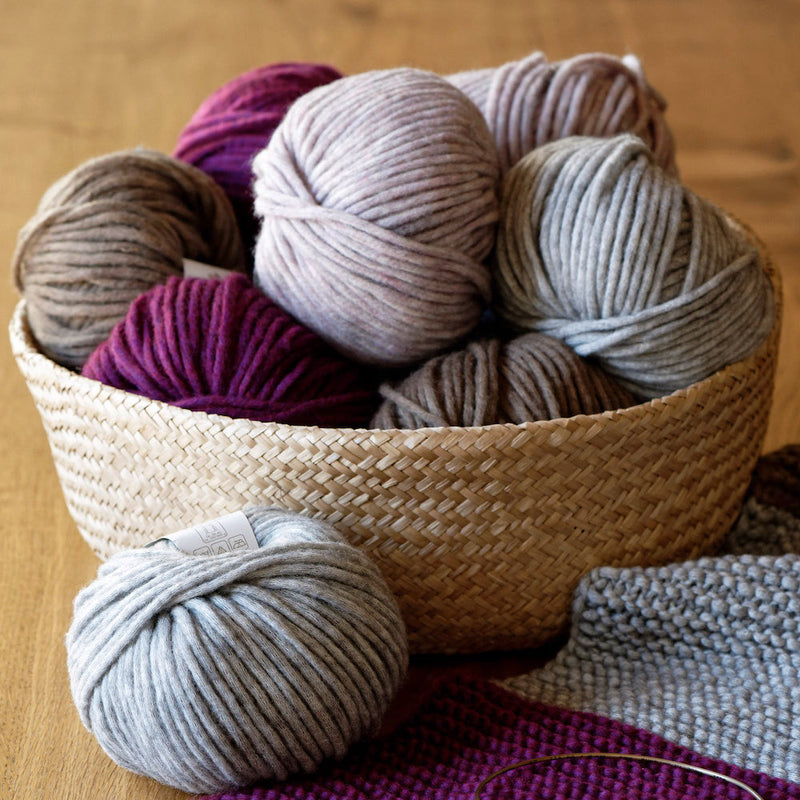 Korb mit mehreren Wollknäueln in verschiedenen Lilatönen und Grautönen, auf Holztisch stehend. Ein Einzelknäuel und gestricktes Stück in Grautönen im Vordergrund.