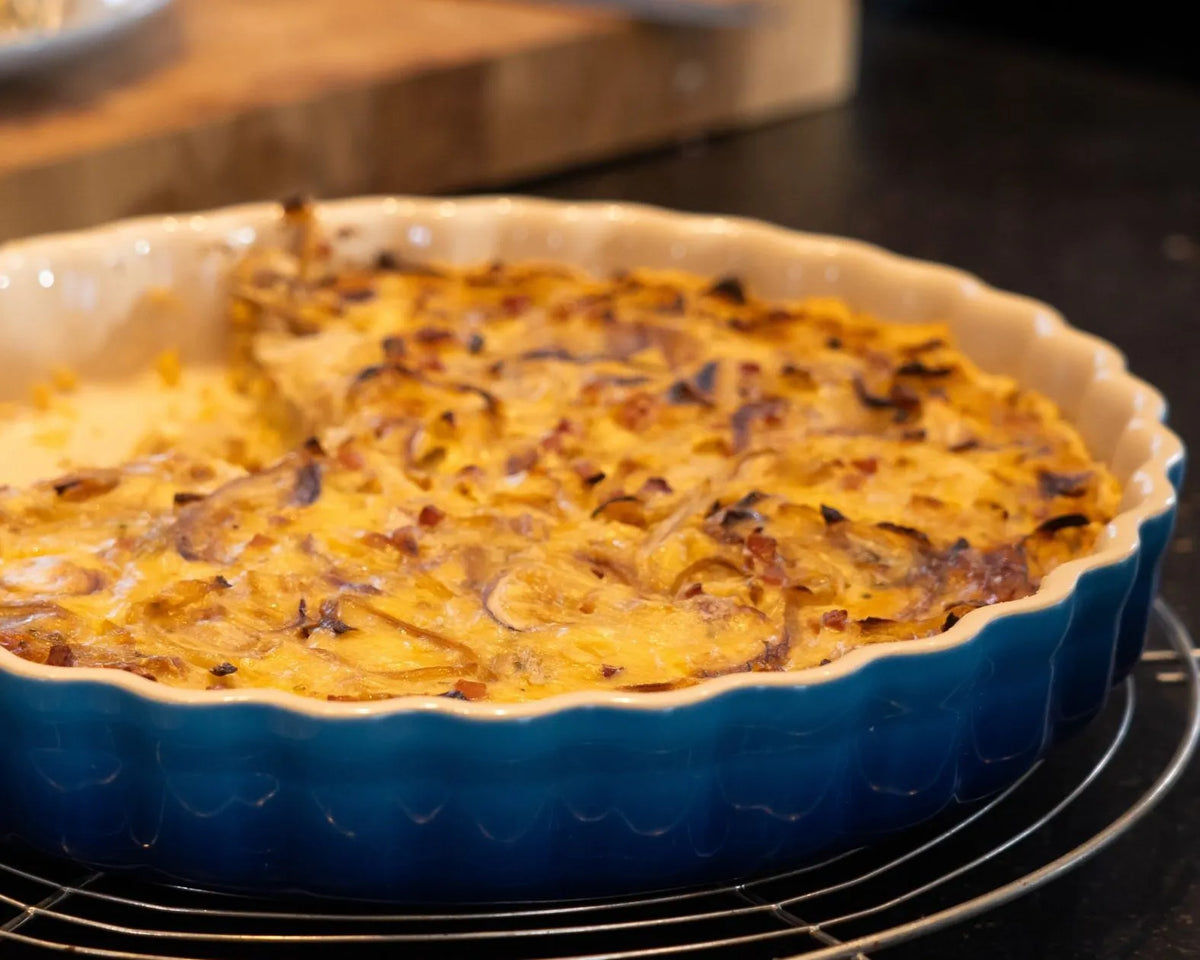 Eine blaue Keramik-Backform auf einem Metallgitter zeigt einen frisch gebackenen Zwiebelkuchen mit goldbraun gebräunten Zwiebeln und Käse überzogen. Die Backform steht auf einer dunklen Küchenarbeitsplatte. Bild verlinkt zum Workshop