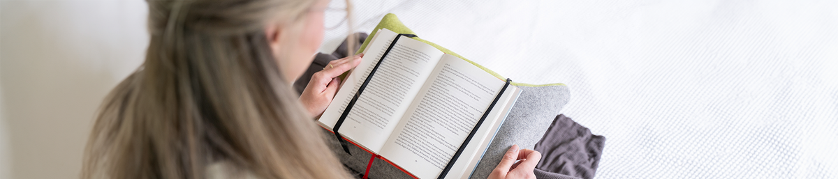 Person sitzt und liest ein Buch. Das Buch ist auf einem speziellen Lesekissen, dass auf dem Schoß der Person liegt, befestigt.