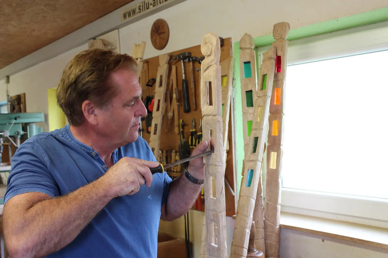 Mann bearbeitet helle Holzskulptur mit rechteckigen Ausschnitten, bunte Glasstücke, Werkzeuge an der Wand, natürliches Licht.