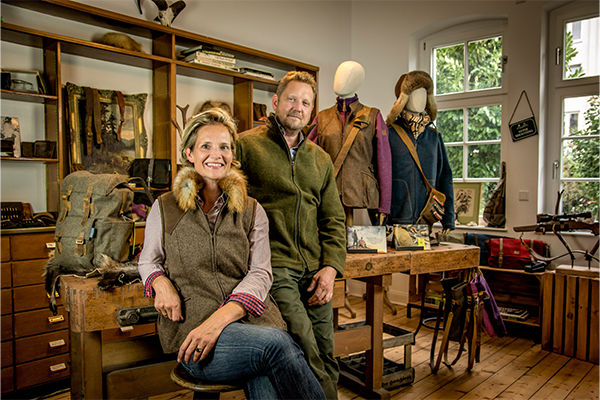 Zwei Personen in rustikalem Atelier, warme Brauntöne, Holzmöbel, Mode mit Fellkragen, Jacken auf Schaufensterpuppen.