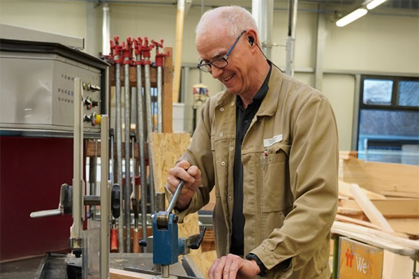 Eine ältere Person mit Brille arbeitet an einer Maschine in einer Werkstatt. Sie trägt eine beige Jacke und hat Ohrstöpsel zum Gehörschutz. Im Hintergrund sind Holzmaterialien und Werkzeuge zu sehen. Bild verlinkt zum Herstellerportrait.