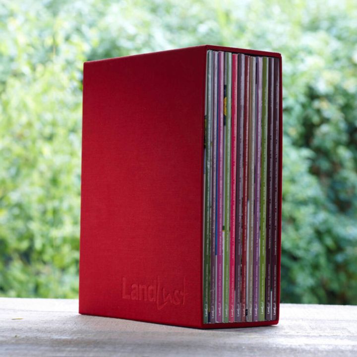Ein roter Zeitschriftensammler mit der Pr&auml;gung "Landlust" steht aufrecht. In ihm sind diverse Zeitschriften sichtbar aufgereiht. Der Hintergrund zeigt unscharfes Gr&uuml;n, m&ouml;glicherweise Pflanzen.