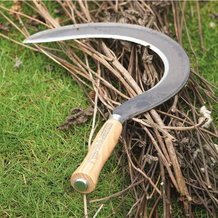 Eine Sichel mit hölzernem Griff und gebogener Metallklinge liegt auf einem Bündel Äste auf grünem Gras.
