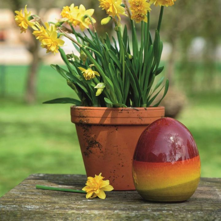 Ein Terrakotta-Topf mit gelben Narzissen steht auf einem Holztisch im Freien. Daneben liegt eine einzelne gelbe Bl&uuml;te und ein gro&szlig;es, buntes, gl&auml;nzendes Osterei mit Farbverlauf von Rot zu Gelb.