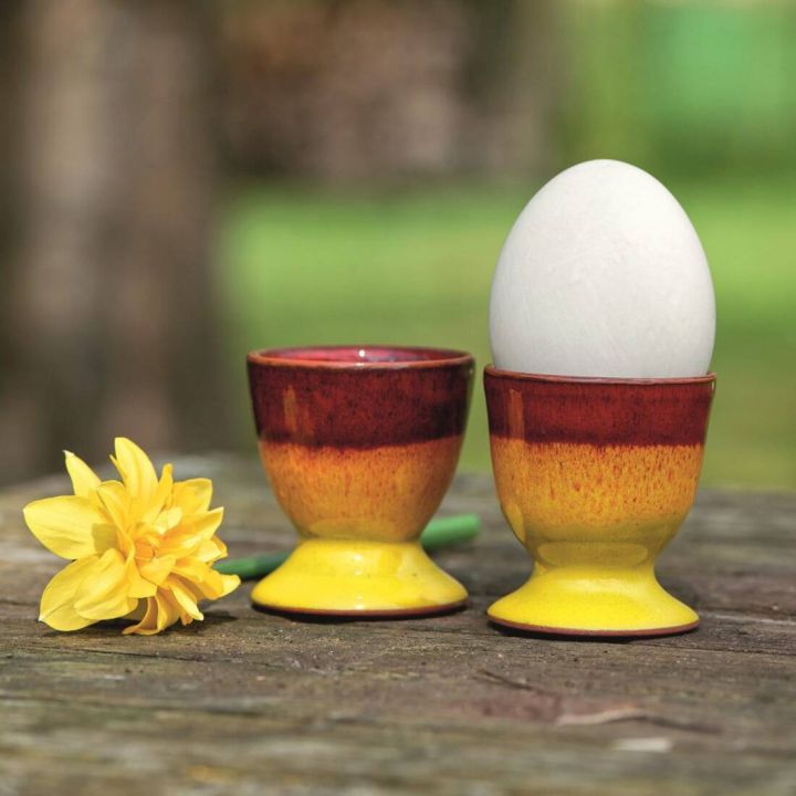 Zwei gelb-orange Keramikeierbecher auf Holztisch im Freien, einer mit weißem Ei gefüllt. Daneben liegt eine gelbe Narzisse. Verschwommener grüner Hintergrund deutet auf einen Garten hin.