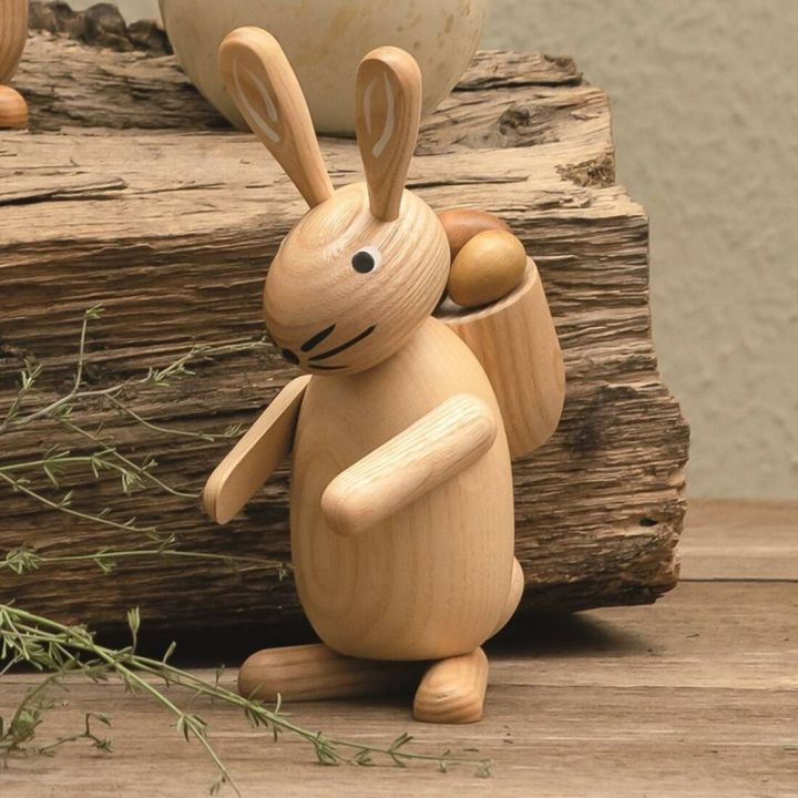 Holzfigur eines stehenden Hasen mit langen Ohren, schwarzem Gesichtsausdruck und beweglichen Armen, trägt auf dem Rücken einen Korb mit zwei Eiern; steht auf Holzuntergrund vor rustikalem Holzblock.