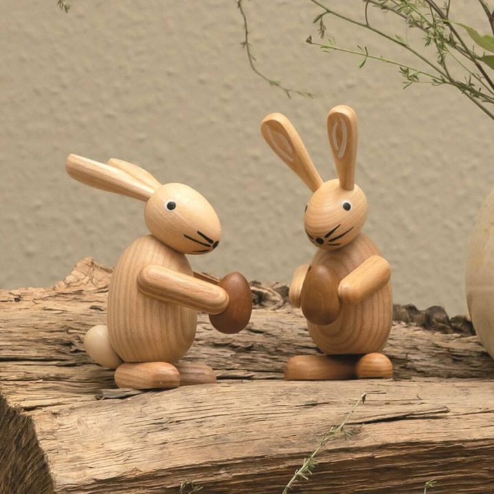 Zwei Hasenfiguren aus Holz, jeweils eine Nuss haltend, stehen auf einem dekorativen Holzstück. Ihre Körper sind hellbraun, mit dunklen Details auf Gesicht und Ohren. Im Hintergrund ist eine strukturierte, beige Wand sichtbar.