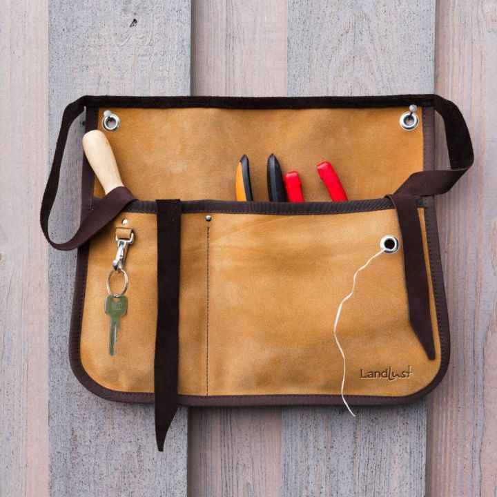 Eine braune Lederwerkzeugtasche mit mehreren Fächern, hängt an einer Holzplatte. Enthält Werkzeuge wie Schraubenzieher und Zangen. Ein Schlüsselbund mit Schlüssel ist an der linken Seite befestigt. Aufdruck: "Landlust".