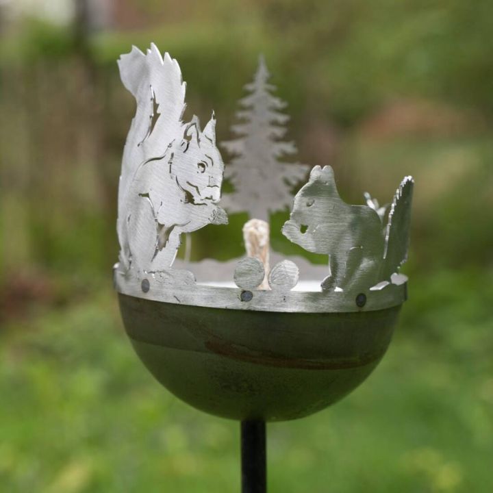 Metallskulptur in Form von zwei ausgestanzten Eichhörnchen und einem Baum auf einer runden Schale, die auf einem Stab montiert ist. Der Hintergrund ist unscharf und zeigt grüne Vegetation.
