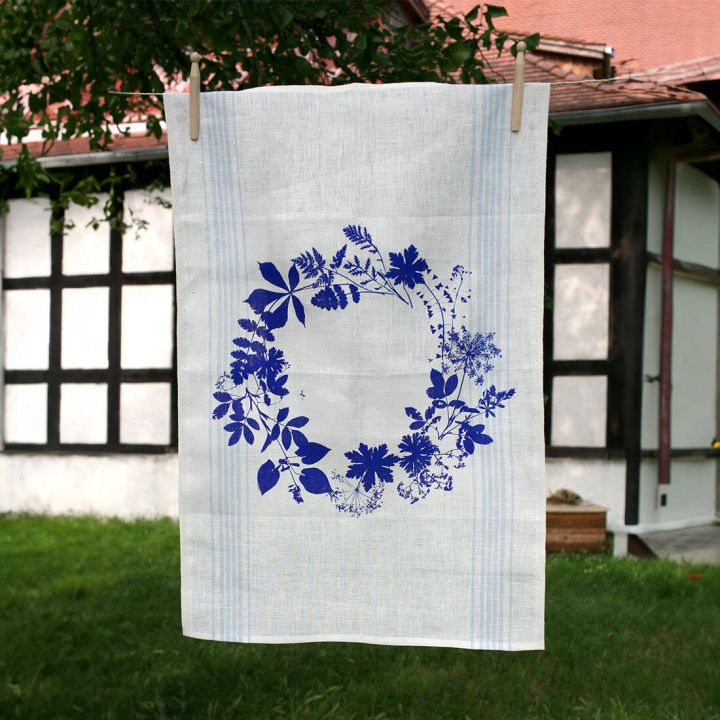 Weißes Geschirrtuch mit einem blauen Blumenkranzmotiv hängt an einer Leine im Freien. Im Hintergrund sind ein Fachwerkgebäude und grüne Bäume sichtbar.