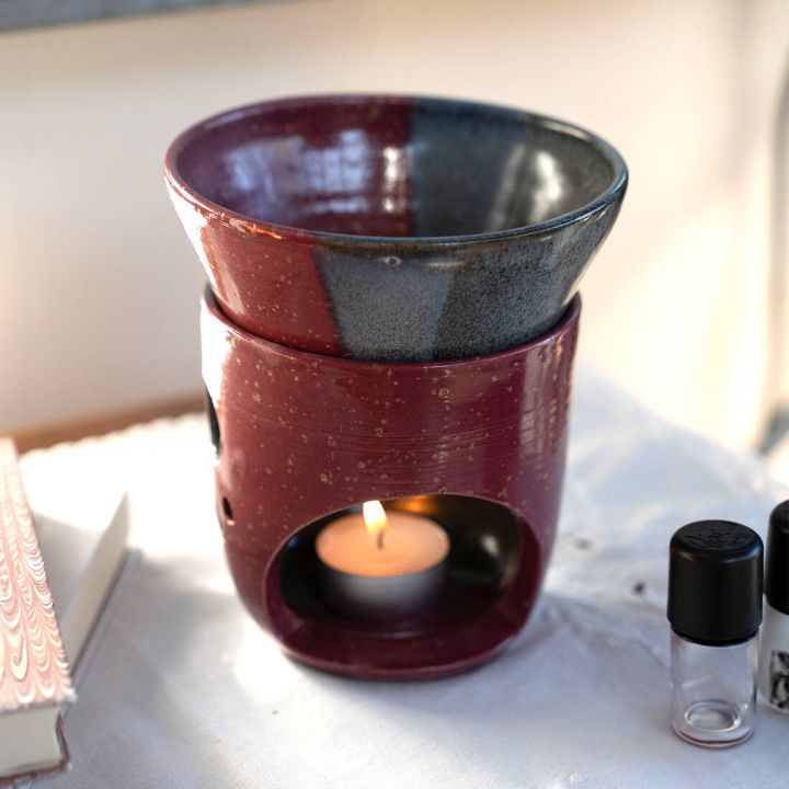 Keramik-&Ouml;lduftlampe in Rot und Grau mit brennendem Teelicht, auf wei&szlig;em Tuch; daneben ein Buch und zwei kleine Fl&auml;schchen.