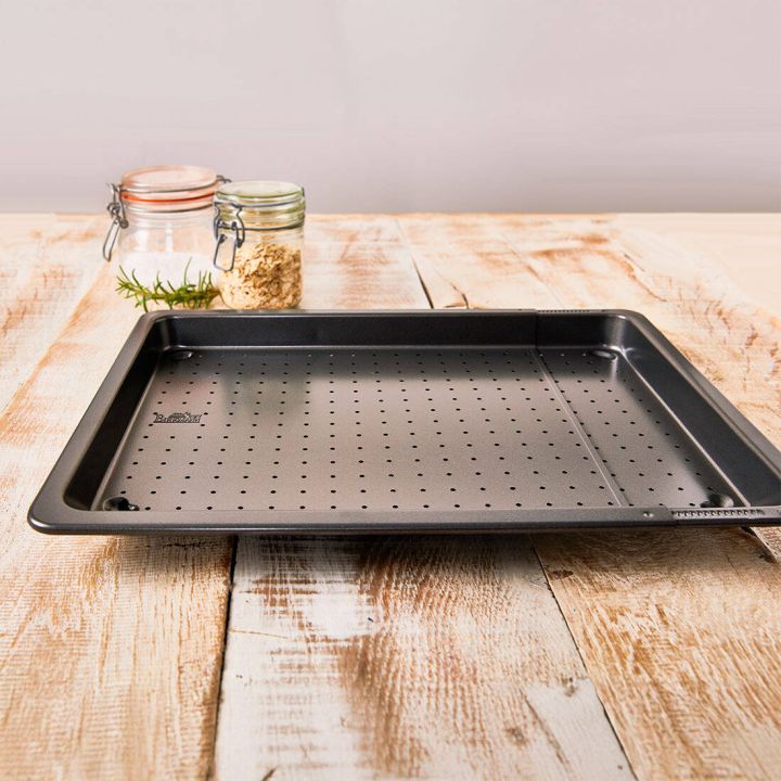 Schwarzes, perforiertes Backblech mit verstellbarer Breite. Das Blech hat kleine Löcher zur besseren Luftzirkulation beim Backen und steht auf einer neutralen Oberfläche.