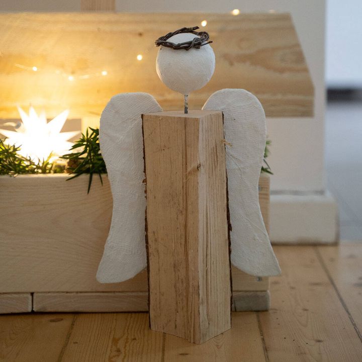 Holzengel-Dekoration mit rechteckigem K&ouml;rperblock, wei&szlig;en Fl&uuml;geln und Kopf aus Kugel mit Dornenkranz. Hintergrund mit Holzstrukturen und weihnachtlicher Beleuchtung, platziert auf Holzboden.