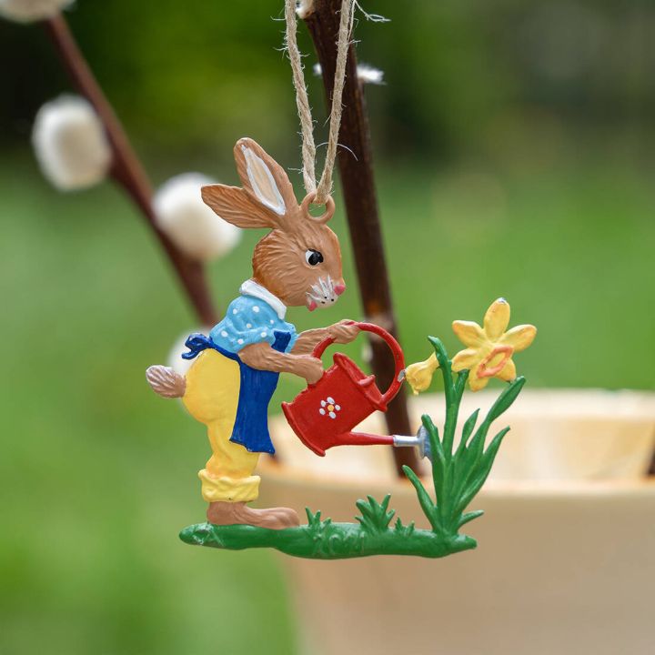 Bunter Osterhaseaufhänger mit blauer Jacke und gelber Hose gießt mit roter Kanne gelbe Blume, unscharfer grüner Hintergrund.