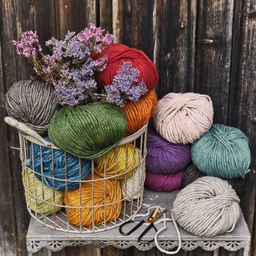 Korb mit bunten Wollknäueln in verschiedenen Farben, ergänzt durch lila Blumen, auf einer Häkeldeckchen-bedeckten Oberfläche. Daneben liegt eine Schere. Hintergrund aus dunklem Holz.