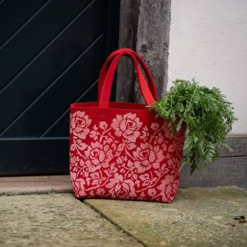 Eine rote Stofftasche mit floralen Mustern, gefüllt mit Karottengrün, steht auf einem Steinboden vor einer dunklen Tür und einer Holzwand.