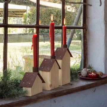 Drei rote Kerzen stehen in Holzhäusern auf einer Fensterbank. Fenster im Hintergrund zeigen grünen Außenbereich. Rechts ein rotes Tablett mit dekorativen Tannenzapfen und einer Teelichtkerze.