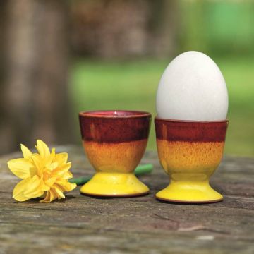 Zwei gelb-orange Keramikeierbecher auf Holztisch im Freien, einer mit weißem Ei gefüllt. Daneben liegt eine gelbe Narzisse. Verschwommener grüner Hintergrund deutet auf einen Garten hin.