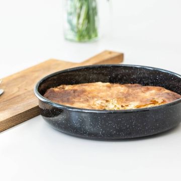 Ein runder Kuchen in einer schwarzen Backform mit weißem Muster auf einer weißen Oberfläche. Im Hintergrund steht ein Holzschneidebrett und eine Vase mit grünen Pflanzen.