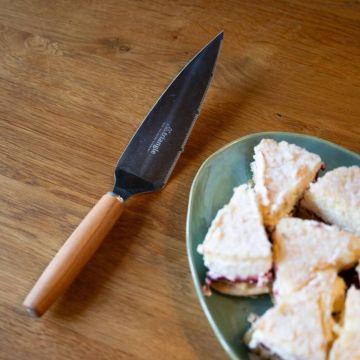 Schneidmesser mit schwarzer Klinge und Holzgriff liegt auf Holztisch neben grünem Teller mit dreieckigen Kuchenstücken, teilweise mit roter Füllung; Messeraufschrift „triangle“.