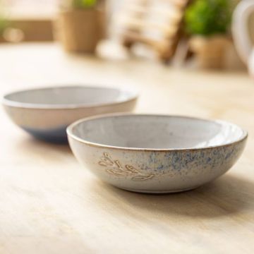 Zwei Keramikschalen auf einem hellen Holztisch. Die vordere Schale hat ein eingeprägtes Pflanzenmuster und blauen Farbakzent. Beide Schalen sind rund und flach, der Hintergrund ist unscharf mit Pflanzen.