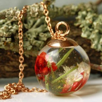 Runder Anhänger mit goldener Kette, enthält getrocknete rote und weiße Blüten sowie grüne Gräser, eingebettet in klaren Harz. Der Hintergrund zeigt unscharfe, natürliche Texturen von Holz und Moos.