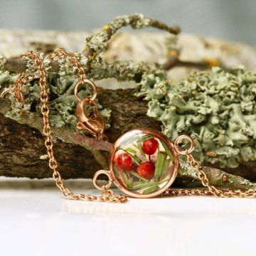 Goldene Halskette mit rundem Anhänger, gefüllt mit roten Beeren und grünen Pflanzen, auf einem moosbewachsenen Ast liegend.