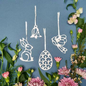 Vier weiße Metall-Osteranhänger – Hase, Ei, Glocke, Vogel – mit filigranen Aussparungen hängen an Satinbändern vor blauem Hintergrund, eingerahmt von rosa Nelken, Chrysanthemen und grünem Blattwerk.