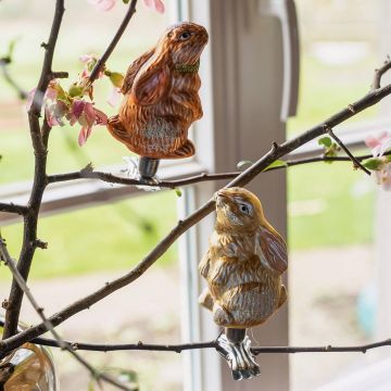 Zwei dekorative Hasenfiguren aus Glas sind auf blühenden Ästen in einem Raum mit großen Fenstern befestigt. Die Hasen sind detailliert gestaltet und funkeln leicht im Licht.