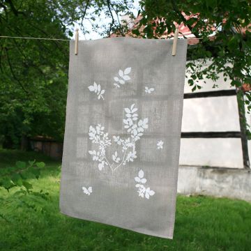 Graues Geschirrtuch mit weißen Blättermotiven hängt an einer Leine im Freien. Im Hintergrund sind ein Fachwerkgebäude und grüne Bäume zu sehen.