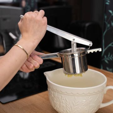 Eine Person presst gekochte Kartoffeln mit einer Edelstahl-Kartoffelpresse in eine beige, verzierten Schüssel. Die Schüssel steht auf einer Holzoberfläche in einer Küche.