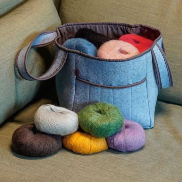 Ein blauer Stoffkorb, gefüllt mit bunten Garnknäueln, steht auf einem Sofa. Vor dem Korb liegen weitere Garnknäuel in verschiedenen Farben wie Braun, Grau, Gelb, Grün und Lila.