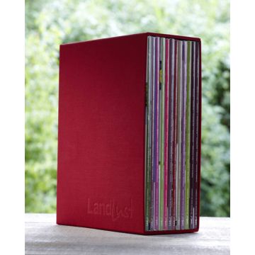 Ein roter Zeitschriftensammler mit der Prägung "Landlust" steht aufrecht. In ihm sind diverse Zeitschriften sichtbar aufgereiht. Der Hintergrund zeigt unscharfes Grün, möglicherweise Pflanzen.