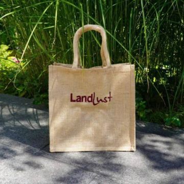 Naturfarbene Jutetasche mit zwei kurzen Henkeln, stehend auf grauem Steinboden vor hohen grünen Gräsern; mittig bedruckt mit dem roten Schriftzug „Landlust“.