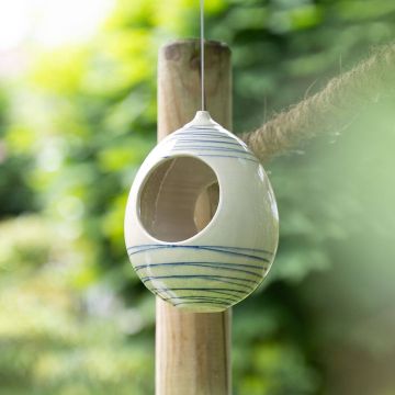 Keramisches Vogelfutterhaus in ovaler Form, weiß mit blauen Streifen, hängt an einem dünnen Seil an einem Baum. Umgebung mit unscharfem grünem Laub und einem weiteren Pfosten im Hintergrund.
