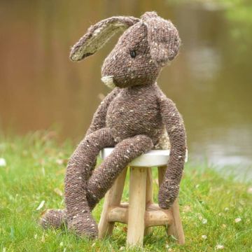 Gestrickter Hase aus brauner Wolle mit weißen Ohren sitzt auf grünem Gras vor unscharfem Hintergrund.