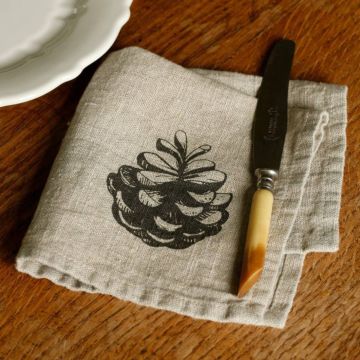 Ein weißer Teller mit gewelltem Rand liegt auf einem Holztisch neben einer weißen Serviette mit aufgedrucktem Tannenzapfen-Motiv. Auf der Serviette liegt ein silberner Löffel.