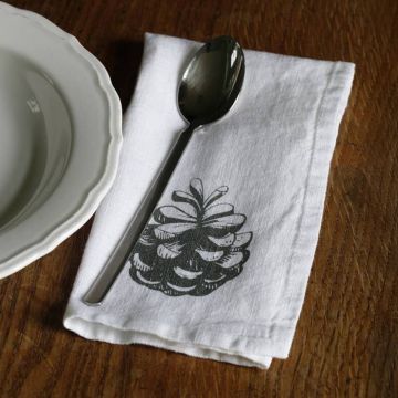 Ein weißer Teller mit gewelltem Rand liegt auf einem Holztisch neben einer weißen Serviette mit aufgedrucktem Tannenzapfen-Motiv. Auf der Serviette liegt ein silberner Löffel.