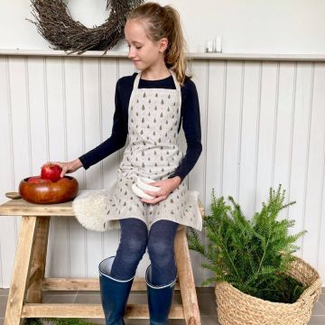 Mädchen im schwarzen Langarmshirt und jeansblauen Leggings, trägt eine beige Schürze mit Baum-Muster. Es sitzt auf einer Holzbank mit einem Wollkissen, hält weiße Schalen, greift nach roten Äpfeln in einer Holzschale.