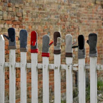 Verschiedene bunte Fäustlinge hängen aufrecht auf einem weißen Holzzaun. Der Hintergrund zeigt eine gemauerte Ziegelwand. Die Handschuhe variieren in Farben und Mustern.