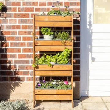 Vertikaler Holzgarten mit vier Ebenen, gefüllt mit verschiedenen Pflanzen und Kräutern, vor einer Ziegelwand. Der Pflanzenständer enthält Basilikum, Petersilie, Thymian und blühende Pflanzen. Teilweise beschattete Fläche neben einer weißen Tür.
