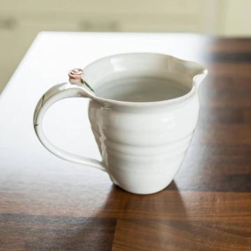Weiße Keramikkanne mit gewellter Oberfläche und kleinem Rosenmotiv am Griff, gefüllt mit Wasser, steht auf einem Holztisch.